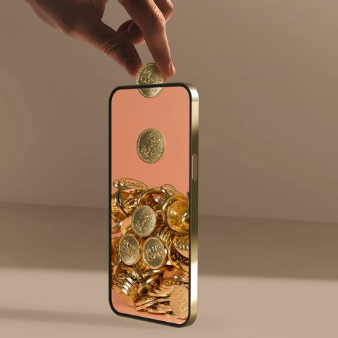 Eine Hand legt eine Silbermünze in ein Smartphone. Auf dem Bildschirm stapeln sich Goldmünzen. Der Hintergrund ist neutral beige. Das Bild suggeriert digitales Sparen oder Investieren.