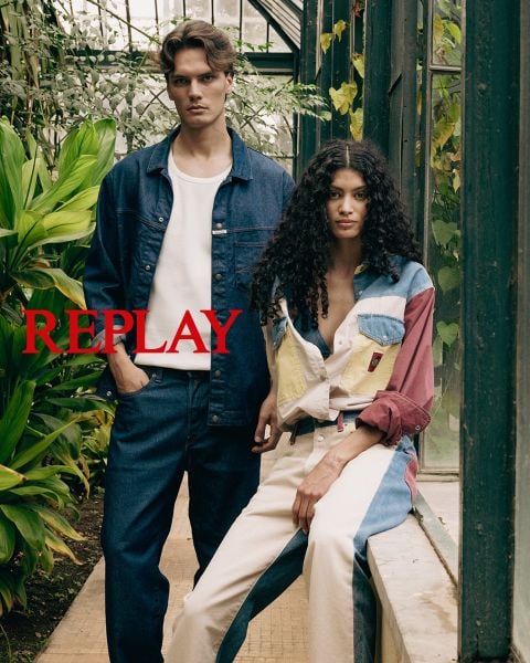 Ein Mann und eine Frau sitzen in dunkler, stilvoller Kleidung auf einer Holztreppe in einem Innenraum. Das Wort "REPLAY" ist in der Mitte des Bildes in roten Großbuchstaben prominent eingeblendet.