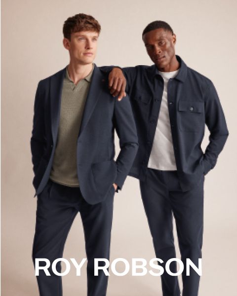 Ein Mann steht im Freien vor verschneiten Bergen und trägt einen grauen Hahnentrittmantel, einen grauen Pullover und eine graue Hose. Der Text "ROY ROBSON" erscheint am unteren Rand des Bildes.