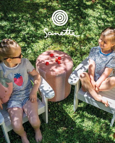 Vier lächelnde Kinder stehen auf einer Wiese und halten Stangen, zwischen denen bunte Stoffstreifen gespannt sind. Zwei Alpakas sind in der Nähe, und im Hintergrund sind ein wolkenverhangener Himmel und Bäume zu sehen. Oben steht "Sanetta" geschrieben.