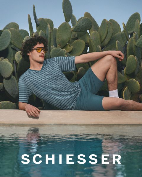 Eine Frau und ein Mann in hellgrauer Loungewear sitzen dicht nebeneinander auf einem Teppich und lächeln sanft in die Kamera. Der Markenname "SCHIESSER" ist im unteren Teil des Bildes zu sehen.