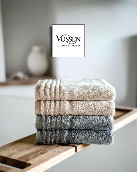 Eine Frau mit kurzen blonden Haaren, eingewickelt in ein graues Handtuch, sitzt auf einer hellen Couch und blickt in die Kamera. In der unteren linken Ecke ist das Vossen-Logo zu sehen.