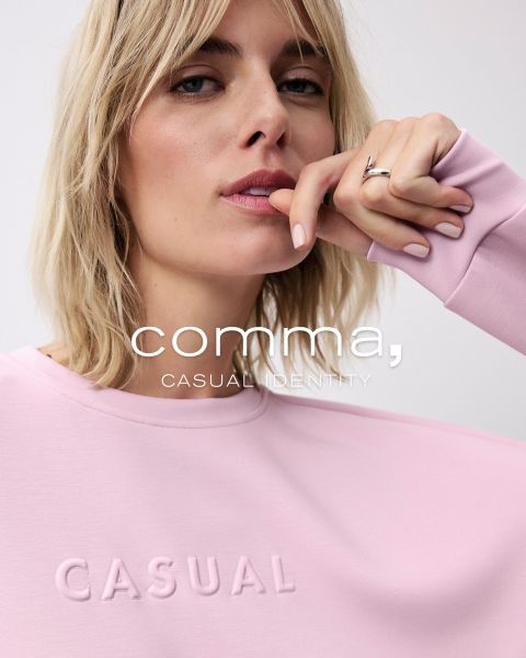 Eine Person mit kurzen blonden Haaren trägt einen hellrosa Pullover, auf dem das Wort „CASUAL“ eingeprägt ist. Darüber ist der Text „Komma, CASUAL IDENTITY“ gelegt. Die Person legt ihre Hand sanft auf den Pullover in der Nähe ihres Kinns.