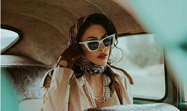 Eine elegante Frau mit Kopftuch, Sonnenbrille, Handschuhen und mehreren Perlenketten sitzt auf dem Rücksitz eines Oldtimers und schaut mit elegantem Blick aus dem Fenster.