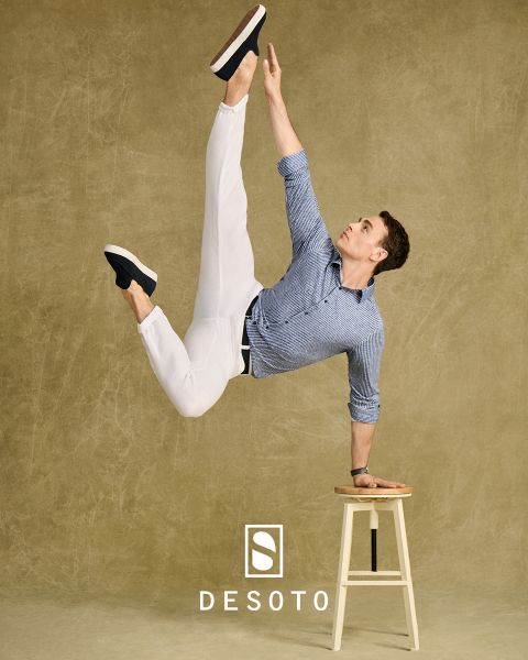Ein Mann in blauem Hemd und weißer Hose führt eine akrobatische Pose auf einem Hocker aus, wobei er auf einer Hand balanciert und ein Bein nach oben streckt. Der Hintergrund ist eine strukturierte beige Wand mit dem Markennamen „DESOTO“ am unteren Rand.