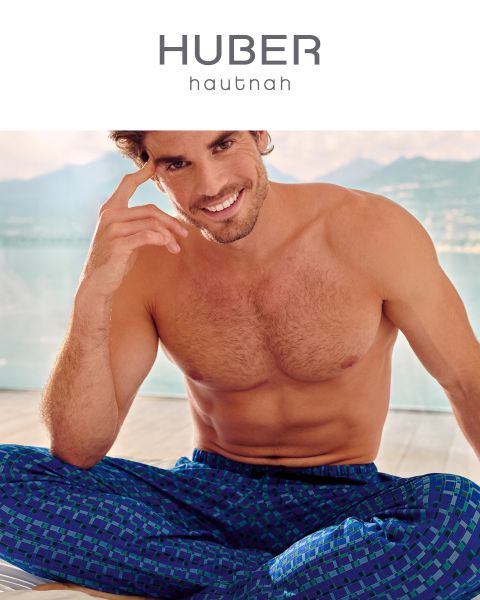 Ein Mann mit kurzen Haaren sitzt im Schneidersitz und lächelt in die Kamera. Er ist oben ohne und trägt eine blau gemusterte Hose. Im Hintergrund ist ein heller, luftiger Raum zu sehen, in der Ferne sind Berge zu sehen. Oben steht der Text „HUBER hautnah“.