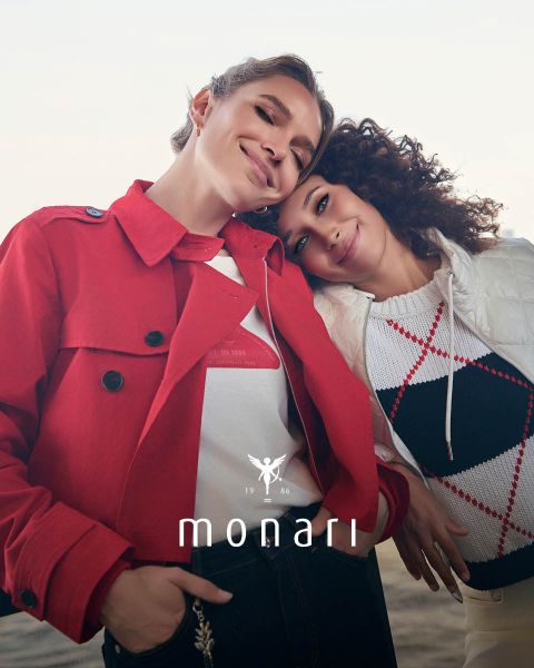 Eine lächelnde Frau, die einen beigen Mantel, ein gestreiftes Hemd, blaue Jeans und einen hellen Schal trägt, steht im Freien vor einem hohen Gebäude. Das Monari-Logo ist am unteren Rand des Bildes zu sehen.