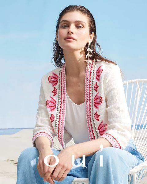 Eine Person sitzt auf einem weißen Stuhl. Sie trägt ein weißes Oberteil und eine offene Jacke mit rosa und roter Blumenstickerei. Sie hat langes, nasses Haar und federförmige Ohrringe. Im Hintergrund sind ein Sandstrand und ein blauer Himmel zu sehen. Unten steht das Wort „oui“.
