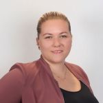 Eine Frau mit heller Haut und blondem, nach hinten gestecktem Haar lächelt leicht, trägt eine lilafarbene Jacke über einem schwarzen Oberteil und eine zarte Halskette und posiert vor einem schlichten, hellen Hintergrund.