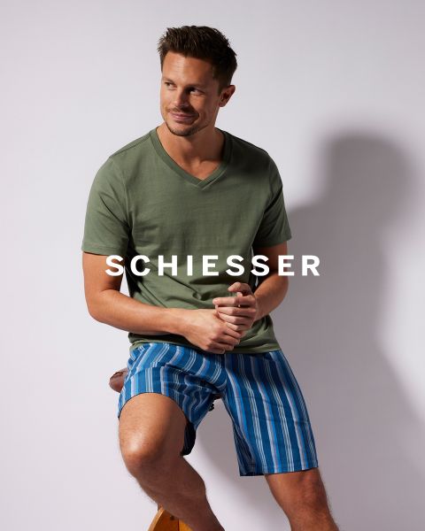 Ein Mann in olivgrünem V-Ausschnitt-T-Shirt und blau gestreiften Shorts sitzt auf einem Holzhocker vor einem einfarbigen Hintergrund. Quer über dem Bild ist das Wort „SCHIESSER“ zu sehen.