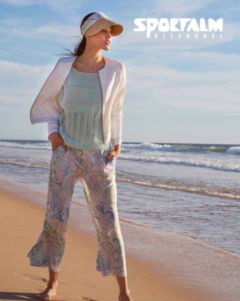 Eine Frau steht an einem Strand. Sie trägt ein hellblaues Strickoberteil, eine weiße Jacke und eine gemusterte Hose. Sie trägt ein Visier und blickt zum Meer. In der oberen rechten Ecke steht „SPORTALM KITZBÜHEL“.