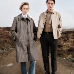 Eine Person in einem Mantel mit Hahnentrittmuster steht neben einer anderen Person, die eine beige Jacke, ein braunes Hemd und dunkle Hosen trägt. Sie befinden sich auf einem Feldweg in einer ländlichen Landschaft. Das Barbour-Logo befindet sich in der oberen linken Ecke.