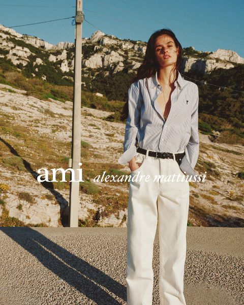 Drei eng beieinander stehende Personen in stilvollen Outfits, eine hält ein Getränk in der Hand. Der Text "ami alexandre mattiussi" ist in weißen Buchstaben über das Bild gelegt. Ihre Gesichter sind nicht zu sehen.
