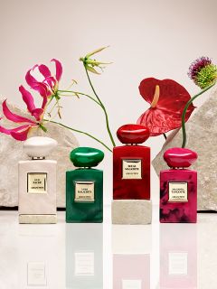 Acht Parfümflakons in verschiedenen Farben, darunter Rot, Grün, Blau, Rosa und Schwarz, sind auf beigen Podesten vor einem hellen Hintergrund angeordnet. Jeder Flakon trägt ein Etikett mit goldener Schrift.