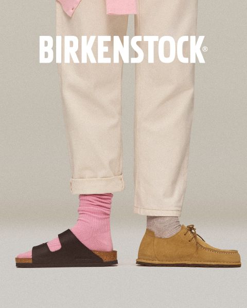 Ein Paar beigefarbene Birkenstock-Clogs mit Schnallenriemen auf einem roten Backsteinboden neben einer roten, geometrisch gefliesten Wand. Das weiße Birkenstock-Logo befindet sich am unteren Rand des Bildes.