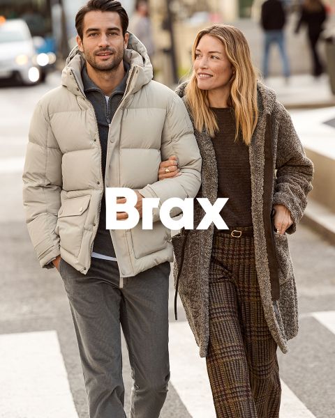 Eine Frau in einem gemusterten Mantel und einem roten Pullover steht neben einem Mann in einem marineblauen Wintermantel und einer beigen Hose. Sie lehnen an einer Steinmauer im Freien. Das Wort "Brax" erscheint am unteren Rand des Bildes.