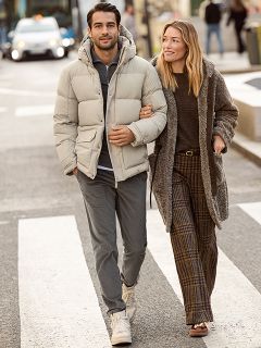 Eine Frau in einem gemusterten Pelzmantel und einem roten Oberteil steht lächelnd neben einem Mann in einer marineblauen Winterjacke und einem beigen Pullover. Sie stehen draußen an einer Steinmauer, und die Frau hält eine braune Handtasche in der Hand.