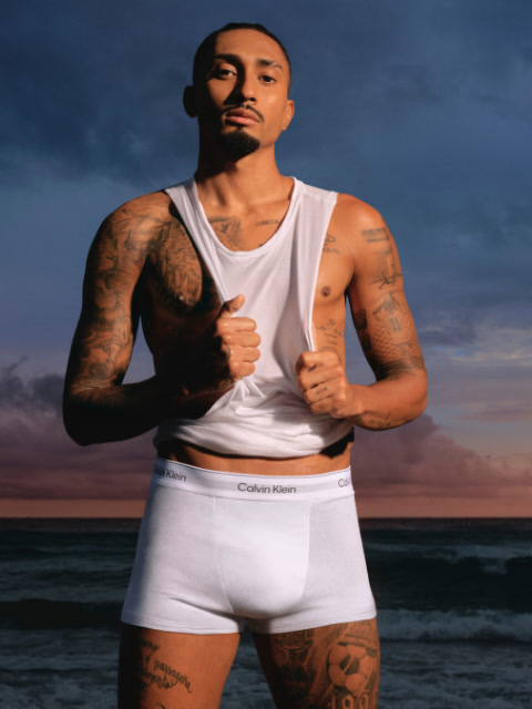 Ein Mann in schwarzer Jacke und Jeans steht in einem Stadion und schaut mit den Händen in den Taschen zur Seite. Im Hintergrund sind Stadionsitze und ein großes Gebäude zu sehen.