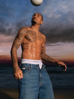 Ein Mann in einer gemusterten Jeansjacke und blauen Jeans steht auf einer Brücke mit Metallgeländer. Im Hintergrund sind ein Fluss und eine weitere Brücke an einem bewölkten Tag zu sehen.