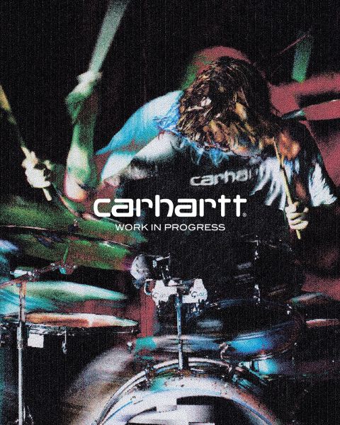 Schwarzweißbild einer Person mit Hut und Jacke, die mit den Händen auf den Knien hockt. Der Text „Carhartt Work In Progress“ ist deutlich in der Mitte zu sehen.