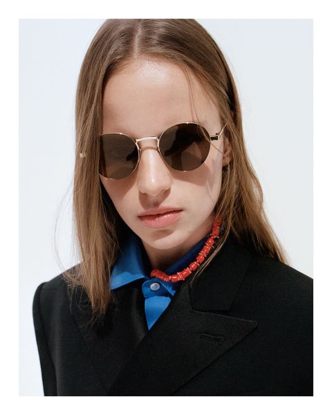 Schwarz-Weiß-Foto einer Person mit kurzem Haar, die eine dunkle Sonnenbrille und ein Hemd mit Kragen unter einer strukturierten Jacke trägt und in einer schwach beleuchteten Umgebung zur Seite schaut.