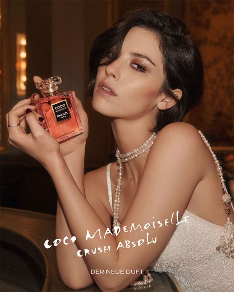 Eine lächelnde Frau in rosafarbener Jacke und schwarzem Rock springt neben einer großen Flasche des Parfums CHANEL Chance Eau Splendide auf weißem Hintergrund.