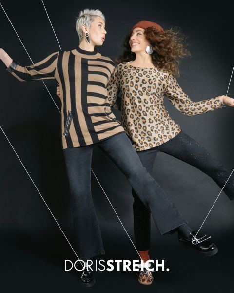 Zwei Frauen posieren selbstbewusst in schwarz-weißen Outfits mit grünen Akzenten, darunter eine Pufferweste, eine Mütze und Schuhe, vor einem dunklen Hintergrund mit dem Text "DORIS STREICH" über ihnen.