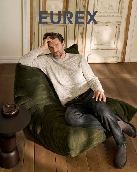 Ein Mann steht im Freien in der Nähe von hohem Gras, trägt einen beigen Pullover, eine marineblaue Pufferweste und eine braune Hose und schaut zur Seite. Das "EUREX by BRAX"-Logo erscheint auf der rechten Seite. Der Himmel ist bewölkt.
