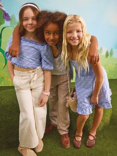 Drei junge Mädchen stehen selbstbewusst zwischen Herbstblättern und tragen metallisch glänzende Outfits. Zwei Mädchen tragen silberne Jacken, und das Mädchen in der Mitte trägt ein mit Pailletten besetztes Oberteil und einen Rock. Ein blaugrüner Hintergrund kontrastiert mit orangefarbenem Herbstlaub.