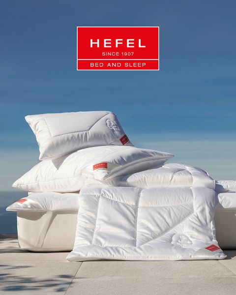 Eine Frau schläft friedlich auf der Seite in einem modernen Bett mit beigem Kissen und weißer Decke. Im Hintergrund sind eine Vase mit Pampasgras und ein Bergblick bei Sonnenuntergang zu sehen. Oben prangt das Logo von „Hefel Bed and Sleep“.