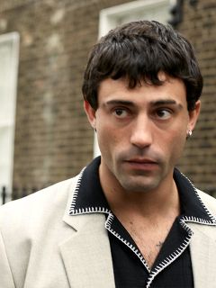 Eine Person im braunen Anzug und mit passender Mütze sitzt auf einer schwarzen Bank vor einem rot-weißen Hintergrund und blickt mit nachdenklicher Miene zur Seite.