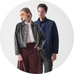 Zwei Personen stehen nebeneinander; die eine trägt eine braune Jacke, die andere eine Sonnenbrille und eine hellbraune Jacke über einem rosa Kleid mit kreisförmigen Mustern.
