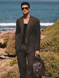 Eine Person in einem formellen schwarzen Anzug steht vor einem Steingeländer mit Bäumen im Hintergrund. Der Himmel ist teilweise bewölkt. Ihr Hemd ist teilweise aufgeknöpft und sie hat einen selbstbewussten Gesichtsausdruck.