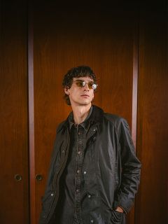 Eine Person mit Sonnenbrille und dunkler Jacke steht vor Holztüren, schaut leicht zur Seite und hat die Hände in den Taschen.
