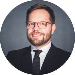 Lächelnder Mann mit Brille, Bart und kurzen Haaren, der einen dunklen Anzug, ein weißes Hemd und eine Krawatte trägt und vor einem grauen Hintergrund posiert.