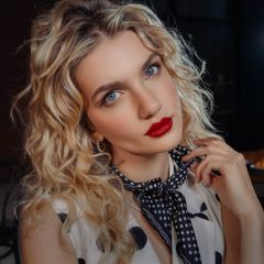 Eine Frau mit lockigem blondem Haar, rotem Lippenstift und einem gepunkteten Schal blickt in die Kamera und hält ihre Hand in den Nacken.