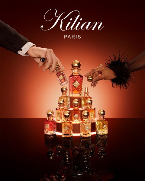Ein Flakon des Parfums Kilian Paris Angels' Share steht neben einem Glas Whiskey mit Eis und einer Garnitur, beides auf einer reflektierenden braunen Fläche mit sanfter Beleuchtung. Oben ist das Kilian Paris Logo zu sehen.
