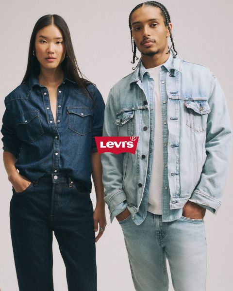 Mann und Frau umarmen sich vor blauem Patchwork-Hintergrund, beide tragen Jeansjacken und Jeans. Über ihnen prangt deutlich ein rotes Levi's-Logo.