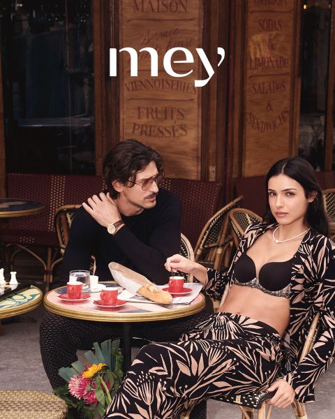 Ein Mann in schwarzen Boxershorts und eine Frau in einem weißen Tanktop und Unterwäsche stehen nebeneinander im Freien an einem Geländer, mit dem Meer und dem bewölkten Himmel im Hintergrund. Das Wort "mey" steht am unteren Rand.