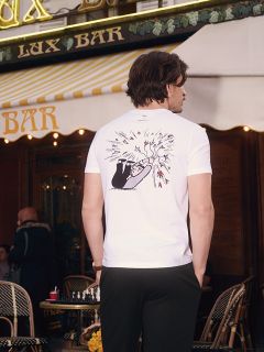 Eine Frau steht in einem Badezimmer und trägt ein lilafarbenes, in der Taille gebundenes Unterhemd, dazu passende Unterwäsche mit hohem Bund und eine durchsichtige Strumpfhose. Sie lehnt an einem Waschbecken vor blassgrünen Wänden und Jalousietüren.