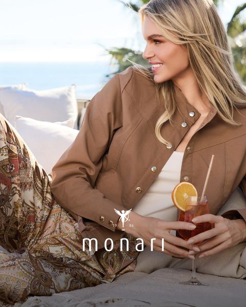 Eine lächelnde Frau, die einen beigen Mantel, ein gestreiftes Hemd, blaue Jeans und einen hellen Schal trägt, steht im Freien vor einem hohen Gebäude. Das Monari-Logo ist am unteren Rand des Bildes zu sehen.