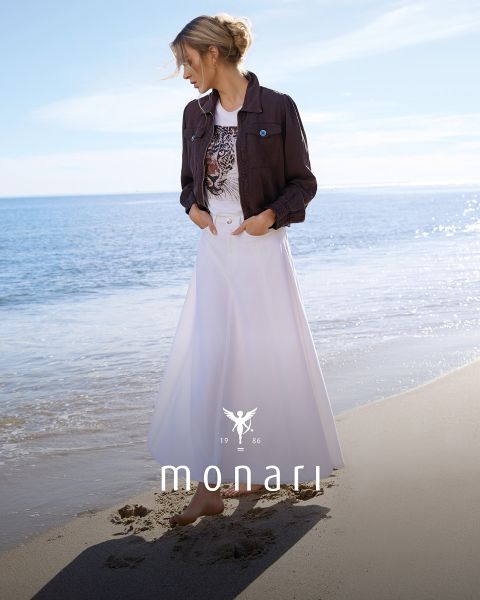 Eine lächelnde Frau, die einen beigen Mantel, ein gestreiftes Hemd, blaue Jeans und einen hellen Schal trägt, steht im Freien vor einem hohen Gebäude. Das Monari-Logo ist am unteren Rand des Bildes zu sehen.