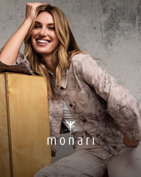 Eine lächelnde Frau, die einen beigen Mantel, ein gestreiftes Hemd, blaue Jeans und einen hellen Schal trägt, steht im Freien vor einem hohen Gebäude. Das Monari-Logo ist am unteren Rand des Bildes zu sehen.