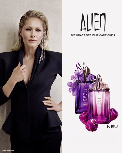 Eine Frau mit strahlend dunkler Haut und glattem schwarzen Haar posiert vor einem violetten Hintergrund. Vor ihr stehen zwei Alien-Parfümflakons von Mugler. Der Text lautet „MUGLER“ und „ALIEN EXTRA INTENSE THE REFILLABLE FRAGRANCES“.