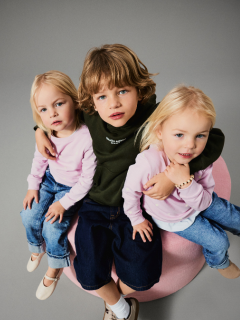 Zwei kleine Kinder stehen Händchen haltend vor einem einfarbig braunen Hintergrund. Ein Kind trägt eine rosafarbene Jacke und einen Rock, das andere eine blaue Jeans und ein blaues Hemd mit der Aufschrift "LIEBE IST KIND". Beide blicken neugierig nach oben.