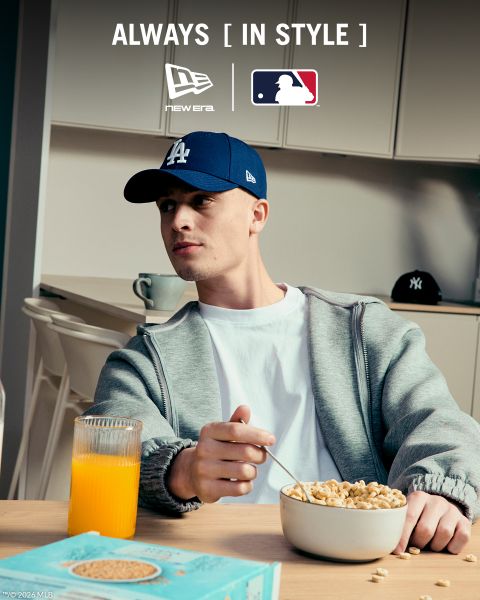 Ein Mann, der einen grauen Trainingsanzug, ein weißes T-Shirt und eine dunkelblaue New York Yankees-Mütze trägt, sitzt auf einer Holzbank und hört mit weißen Kopfhörern Musik. Der Text wirbt für die 9FORTY M-Crown-Kappe von New Era mit dem MLB-Branding.