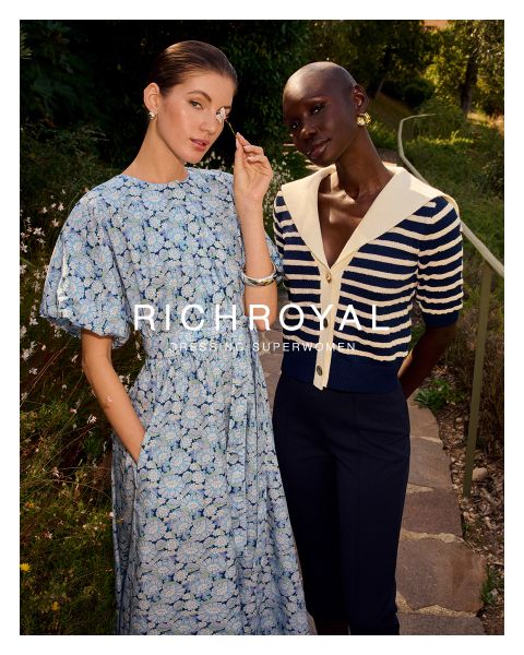 Eine Frau in hellblauem Hemd und grauer Hose hält sich eine Schneekugel vor ein Auge. Sie posiert in einem Raum mit Holzwänden und einem Skiplakat und trägt große Ohrringe. Der Text lautet "RICHROYAL DRESSING SUPERWOMEN".