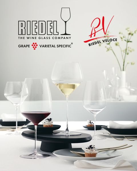 Sechs farbenfrohe Kristallgläser mit aufwendigen Schliffmustern sind auf einer Oberfläche aus dunklem Marmor angeordnet. Darüber prangt das Riedel-Logo mit dem Markennamen und einem Weinglas-Symbol.