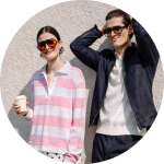 Ein Mann und eine Frau stehen dicht beieinander, beide tragen legere Jacken, vor einer weißen Backsteinmauer und schauen in die Kamera.