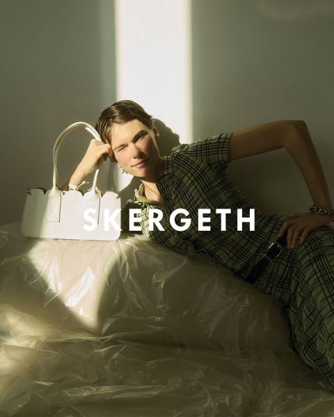 Person in einem schwarzen Blazer über einem gestreiften Hemd und blauen Jeans, die eine schwarze Handtasche hält. Der Text „JULIA SKERGETH“ ist deutlich sichtbar über das Bild gelegt.